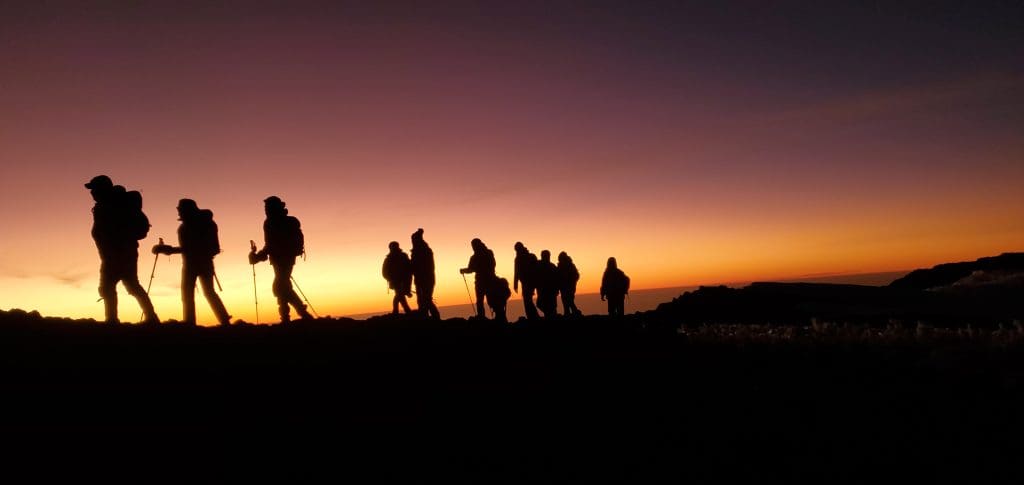 Silhouettes of climbers trekking toward the summit of Mount Kilimanjaro at dawn, with a glowing orange and purple sky in the background.