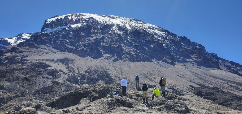 Climbers trekking toward the towering peak of Mount Kilimanjaro, Africa’s tallest mountain, under clear blue skies with the snow-capped summit visible in the background.