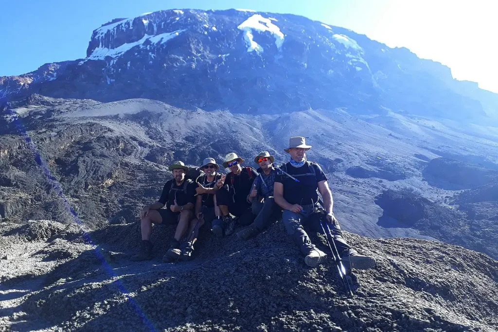 A group of climbers resting on a rocky slope with Mount Kilimanjaro’s snow-capped summit rising majestically in the background.