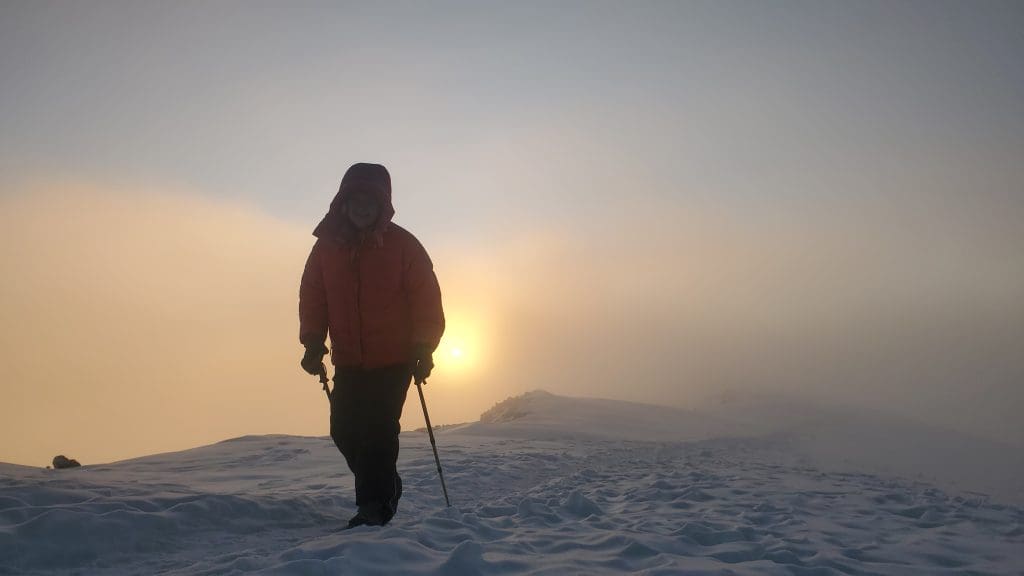 A climber in a red jacket stands on the snowy summit of Mount Kilimanjaro at sunrise, with trekking poles in hand and the golden sun glowing through the mist.