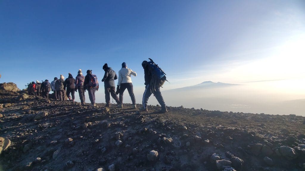 A group of climbers trekking along the rocky summit ridge of Mount Kilimanjaro at sunrise, silhouetted against a clear blue sky with Mount Meru visible in the distance.