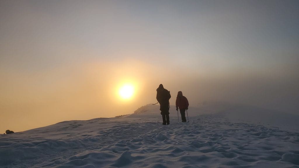 Two climbers walking across snow on Mount Kilimanjaro at sunrise, silhouetted against the golden sky near the summit.