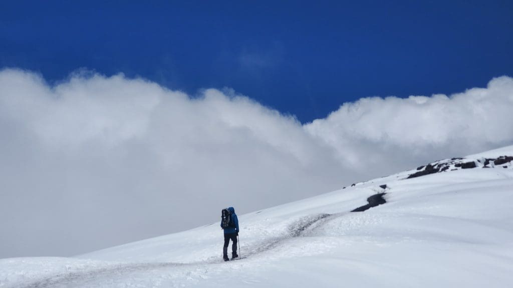 Hiker walking along a snowy trail near the summit of Mount Kilimanjaro under a deep blue sky and clouds.