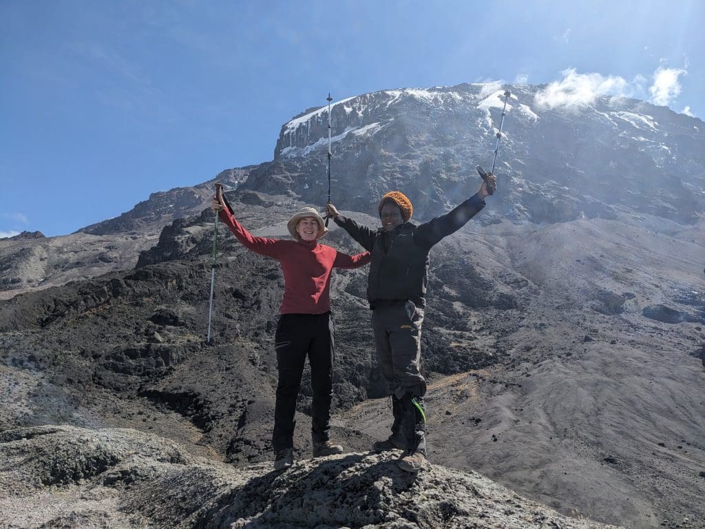 Climber and Kilimanjaro guide celebrating with trekking poles raised, standing on rocky terrain with the snow-capped summit of Mount Kilimanjaro in the background.