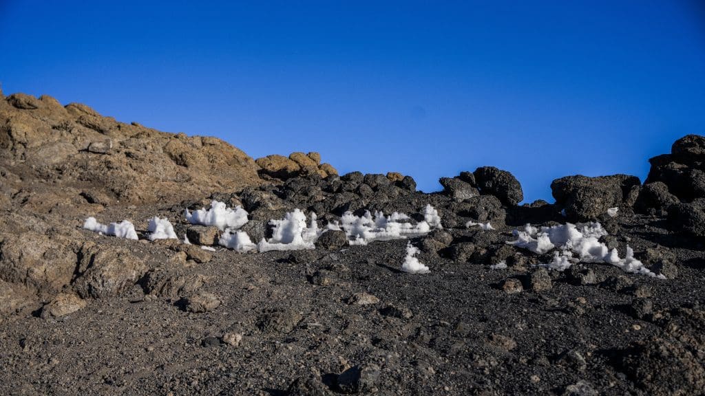 Small patches of snow resting on dark volcanic rocks near the summit of Mount Kilimanjaro under a clear blue sky.