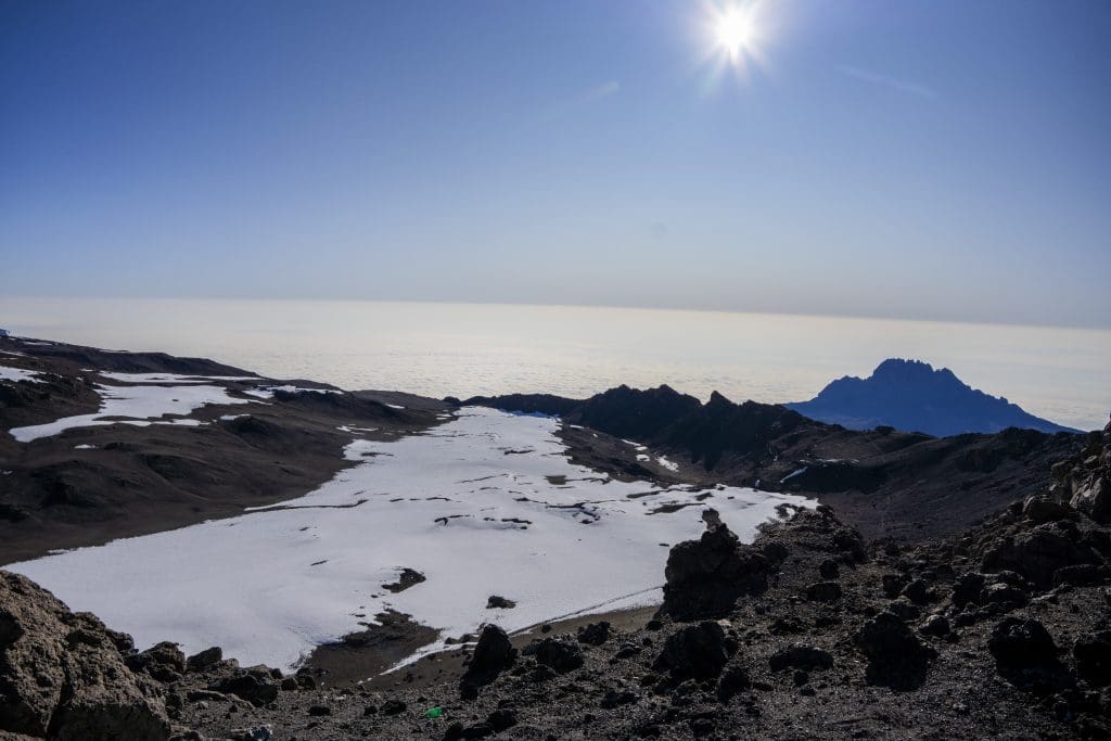 Snow-covered crater landscape on Mount Kilimanjaro with rocky terrain, bright sunlight, and a sea of clouds below.
