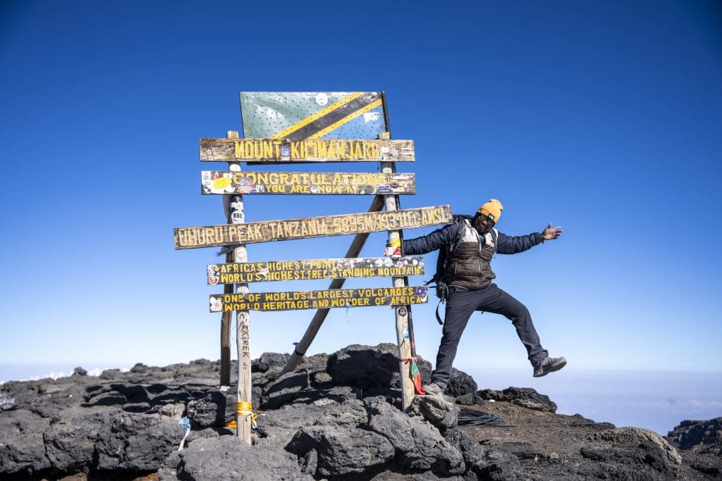 Climber celebrating at the Uhuru Peak sign on Mount Kilimanjaro, Africa’s highest point at 5,895 meters above sea level, under a clear blue sky.